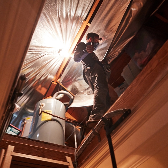 The HeatBusters install a radiant barrier. A technician stands on a wood flooring view from attic door